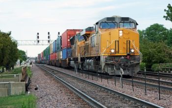 Two yellow locomotives and double stack freight train passing through town