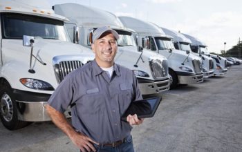 A truck driver is standing in a large parking lot in front of several large trucks.  He is facing the camera, and he is wearing a short-sleeved blue shirt, blue jeans, a brown belt and a gray hat.  He is holding a black tablet with his left hand.  There are six big rigs seen lined up behind him, going from left to right.  The large trucks all look the same, being all white and having silver grills and mirrors.  There are multiple cars seen parked to the right of the trucks.  A cloudy blue sky is seen in the background.